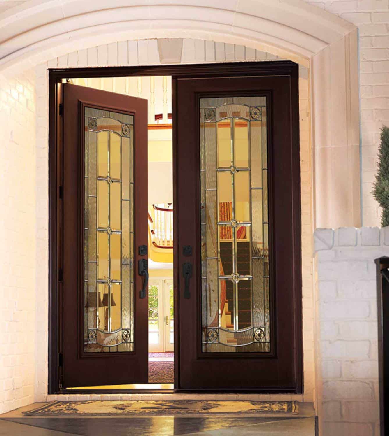 A double-door entrance made of dark wood with intricate stained glass panels stands open, revealing an elegant interior with a warmly lit hallway and spiral staircase in the background. The doorway, framed by light-colored bricks, hints at the quality craftsmanship you'd expect from a top window company in Baton Rouge.