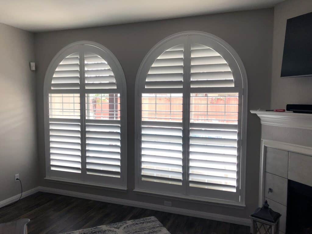 Two large, arched windows with white plantation shutters from a renowned window company Baton Rouge are shown. The partially open windows allow light to filter through. The walls are painted a soft gray, and part of a fireplace with a mounted TV is visible in the room.