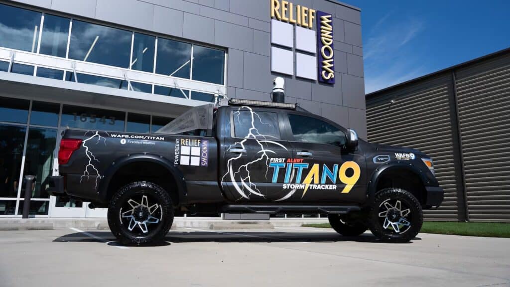 A black pickup truck labeled "First Alert Titan 9 Storm Tracker" is parked in front of a modern building with a "Relief Windows" sign. The Titan 9 features storm-themed graphics and equipment mounted on its roof.