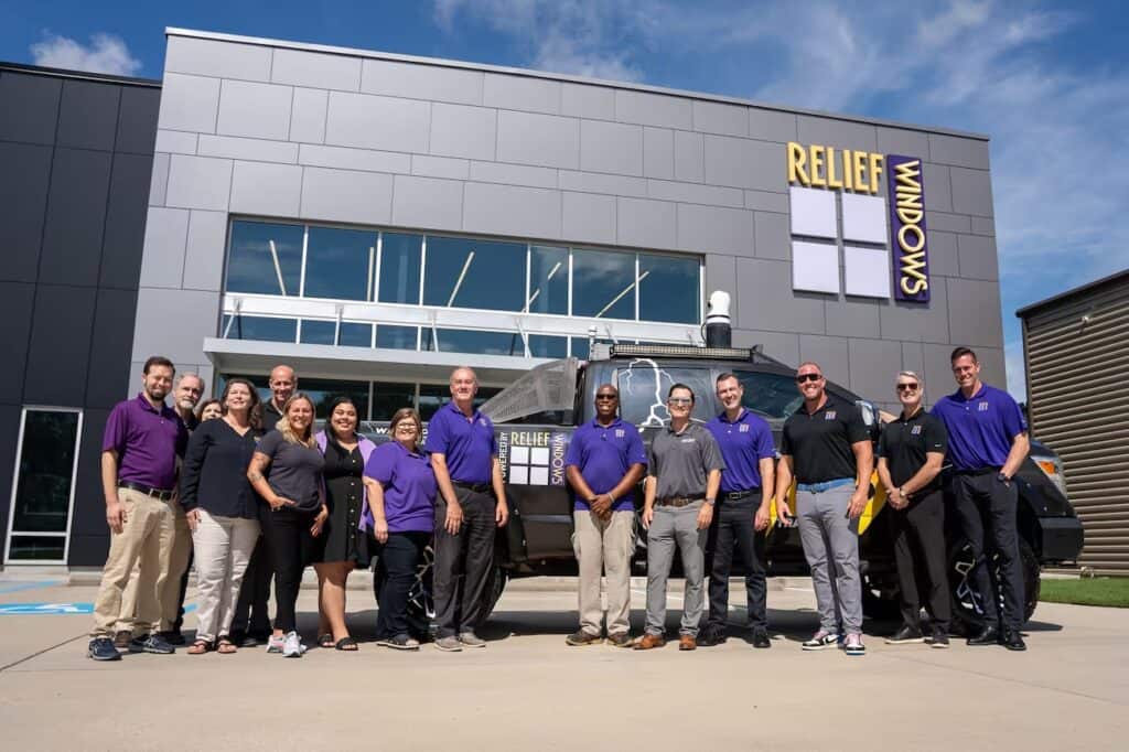 A group of 13 people, wearing casual and business casual attire, stand in front of a modern building with a "Relief Windows" sign. Some members pose with a Titan 9 high-performance vehicle. The background shows a clear, sunny day with blue sky.