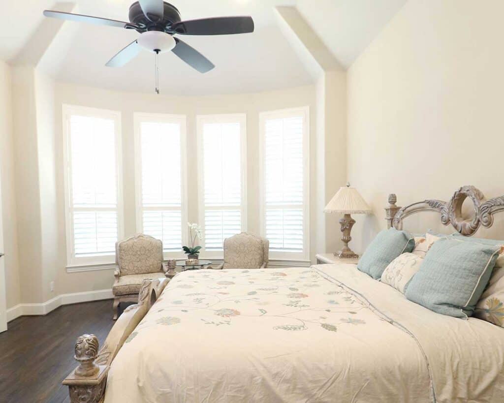 A bright bedroom featuring a large bed with a cushioned headboard, two armchairs by the bay window for an inviting atmosphere, a ceiling fan gently circulating air, and a lamp perched on a decorative nightstand—all enhanced by the quality of Relief Windows Hammond.