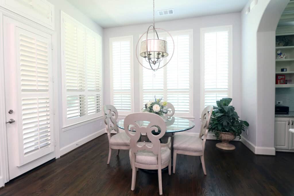 A dining area boasts a round glass table surrounded by four ornate chairs, enlivened by a lush plant and pendant light. Large Relief Windows Lafayette shutters frame the background, adding elegance and charm.