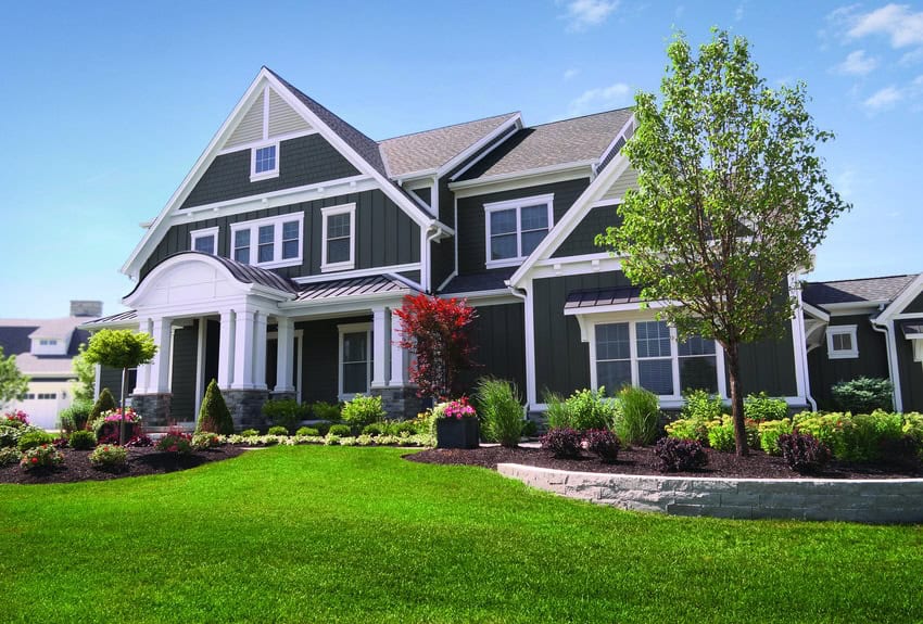 A large two-story house with green siding and white trim stands proudly under a clear blue sky. Enhanced by Relief Windows, it's surrounded by a well-kept lawn and various trees and shrubs, exemplifying the charm of Lafayette living.