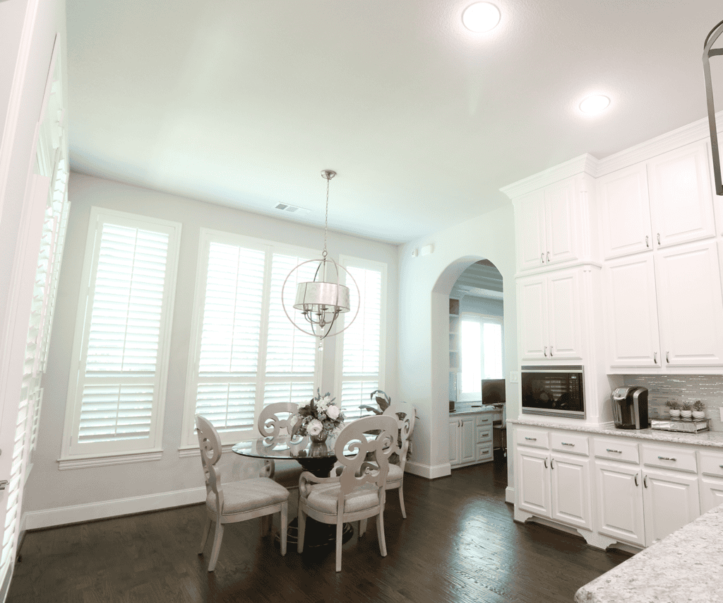 A bright kitchen in Baton Rouge featuring white cabinets, a round dining table with four chairs, and a glistening chandelier. The elegant hardwood flooring complements the large windows with shutters that offer a sense of relief and openness to the space.