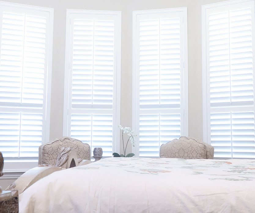 A bedroom with white walls features a bed, two ornate chairs, and four Relief windows by Covington with white shutters, allowing soft light to gently filter into the room.