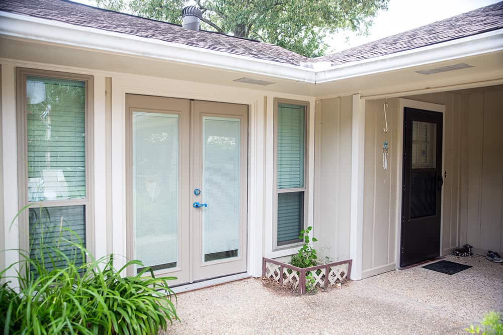 A corner of a house with cream-colored walls, Relief Windows' elegant glass doors, and large windows. There's a plant near the door, adding a touch of greenery to the small tiled patio area.