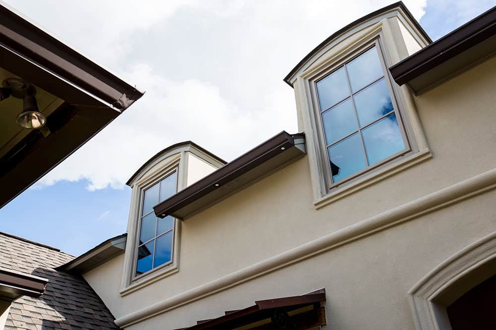 Two dormer windows grace the beige stucco building, offering a serene view of the sky and clouds—a picturesque scene reminiscent of those captured by Relief Windows Slidell.