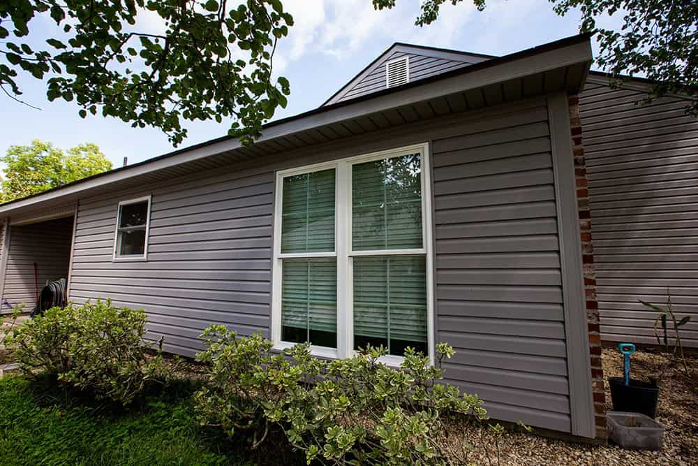 A gray house with siding and large relief windows is surrounded by lush shrubs in Slidell. A small shovel rests near the window, while trees create a serene backdrop.