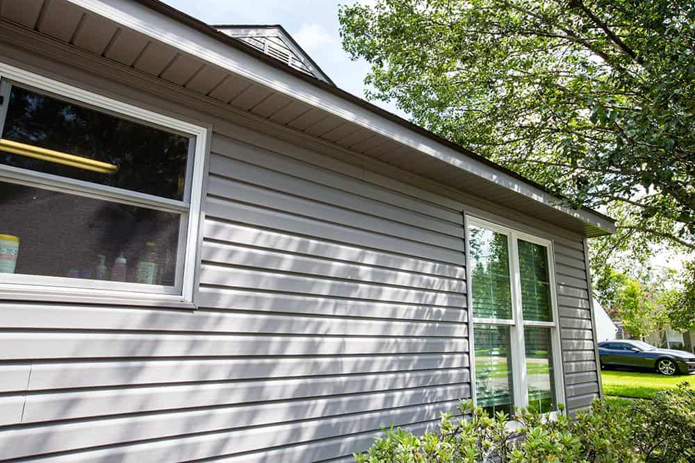 A house with gray siding, Relief Windows blinds, and bushes in front. A tree partially shades the home in Hammond, where a car is visible in the background.