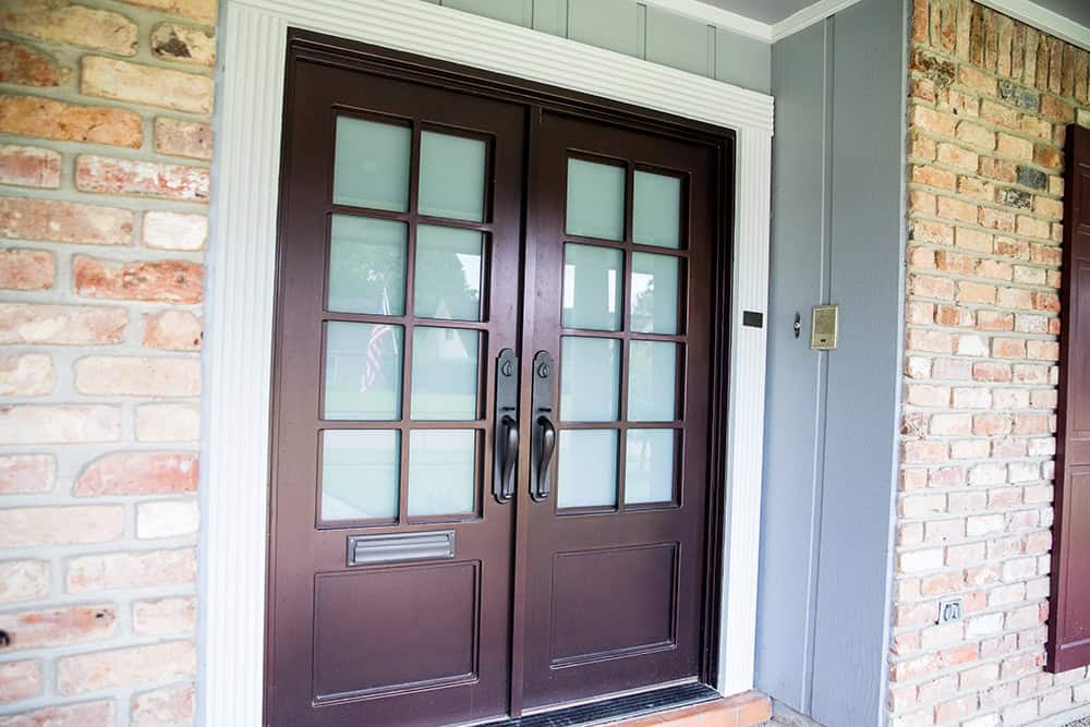 A dark brown double door with glass panes, flanked by brick and gray siding, features a mail slot and two door handles. Crafted by Relief Windows Hammond, it combines durability and elegance seamlessly.