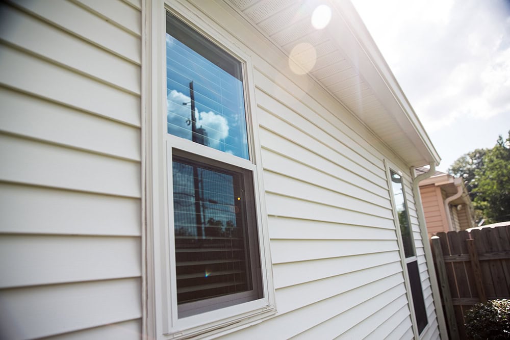 Close-up of a white house exterior, showcasing a window with partially open blinds. Bright sunlight reflects off the siding near Relief Windows in Covington, with a wooden fence completing the scene.