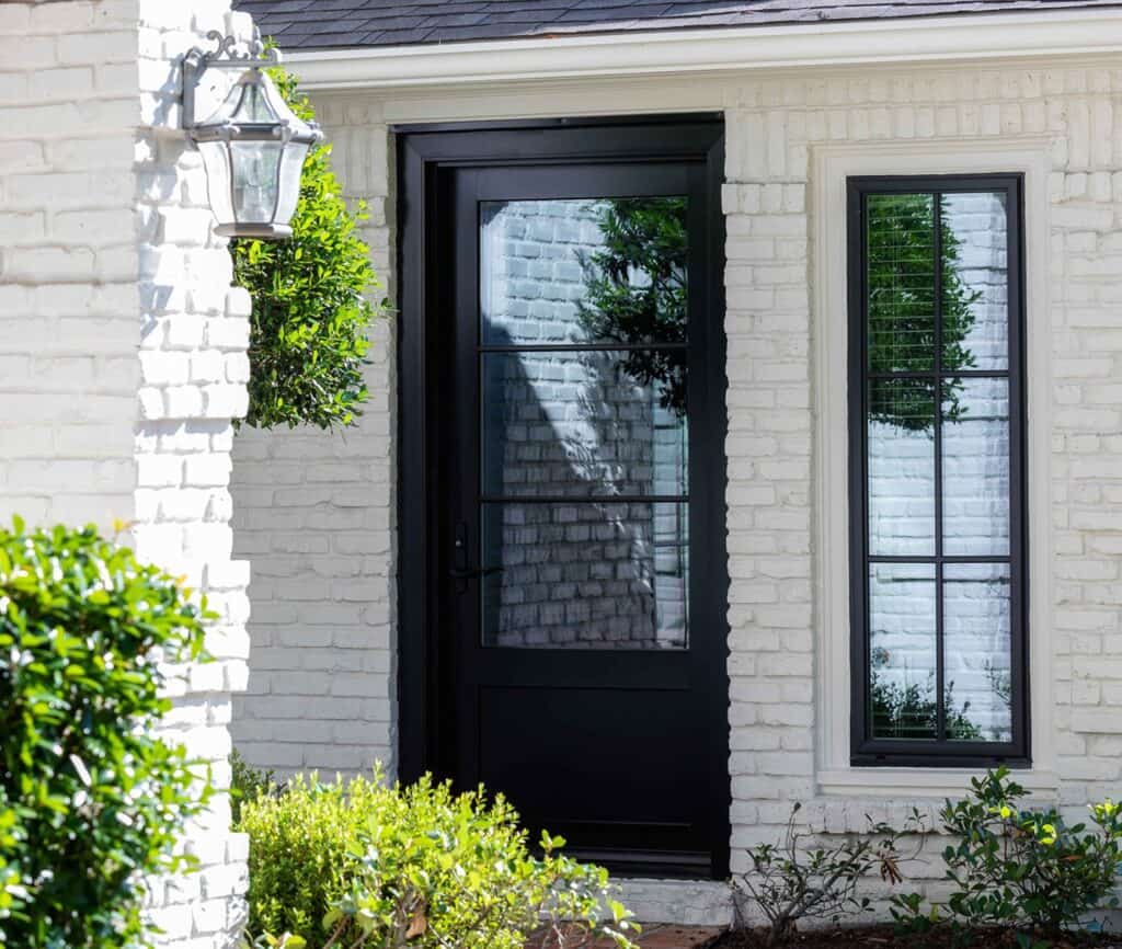 A black framed glass door and window grace the white brick exterior of a house, surrounded by lush green shrubs, offering relief from the Mississippi sun.