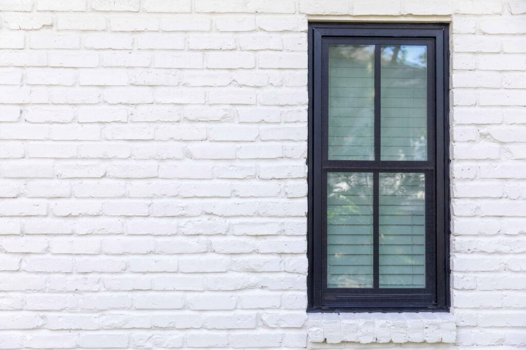 A black-framed window with closed blinds, reminiscent of those from Relief Windows Hammond, is set in a white brick wall.