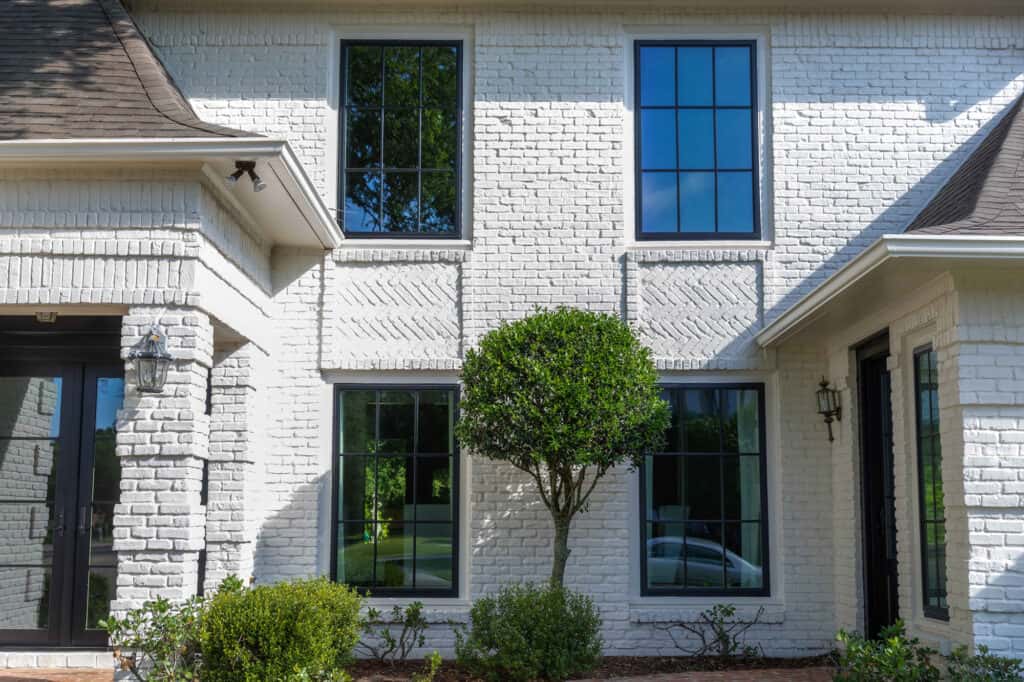 A modern white brick house exterior with large black-framed windows by Relief Windows Mississippi, complemented by a small round tree and neatly trimmed hedges in the front yard.