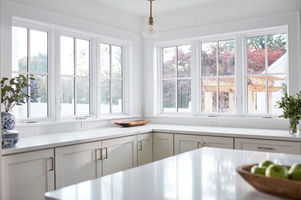 Bright kitchen with white cabinets and countertops, enhanced by Relief Windows Beaumont. Large windows offer abundant natural light, while potted plants add a touch of greenery. A bowl of apples sits invitingly on the counter, completing this charming space.