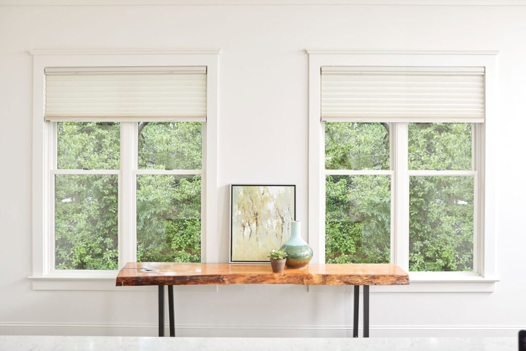 A wooden table sits before two large Relief Windows with white blinds, adorned by a painting, a vase, and a small plant. Green foliage is visible outside.