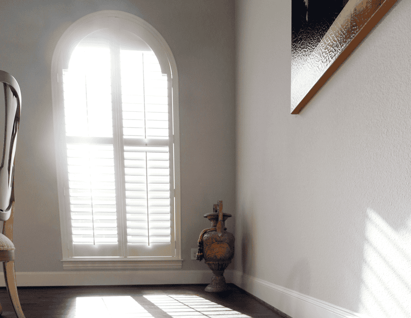 A sunlit room with an arched window featuring Relief Windows Slidell shutters, a decorative jug in the corner, and a partially visible chair and painting on the wall.